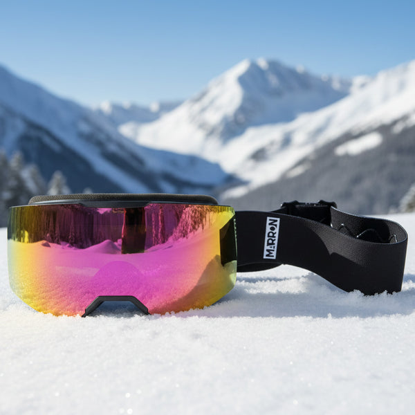 MARRON Skibrille THE ONE mit gold-pink verspiegelter Linse liegt im Schnee, im Hintergrund unscharfe Bergkulisse.